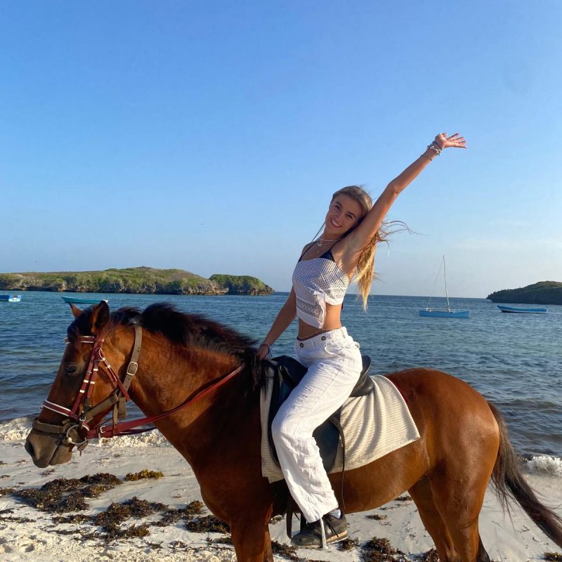 Young girl on a horse on a beach with her arm in the air and looking happy.